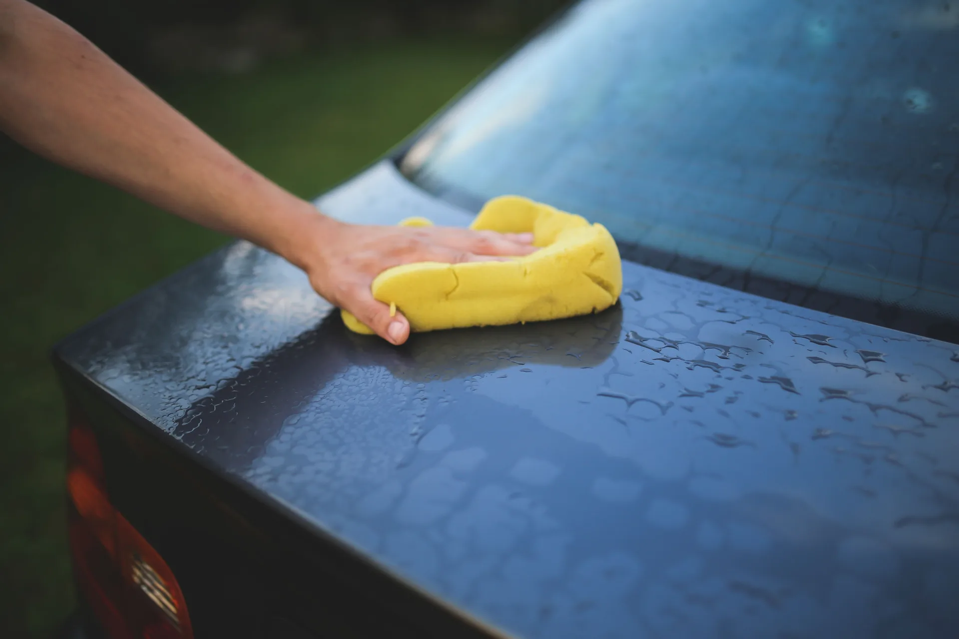 A hand drawing a microfibre cloth across the wet rear panel of a dark car, water still beading on the paint